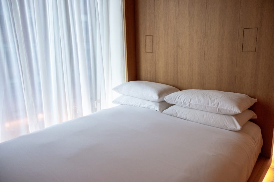 A made bed with fresh white pillows and linen beside a window with white curtains