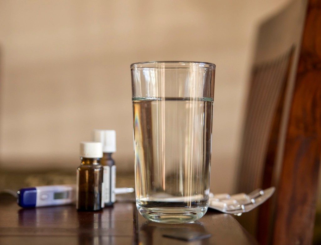 A glass of water beside small medicine bottles and a blister strip on a table