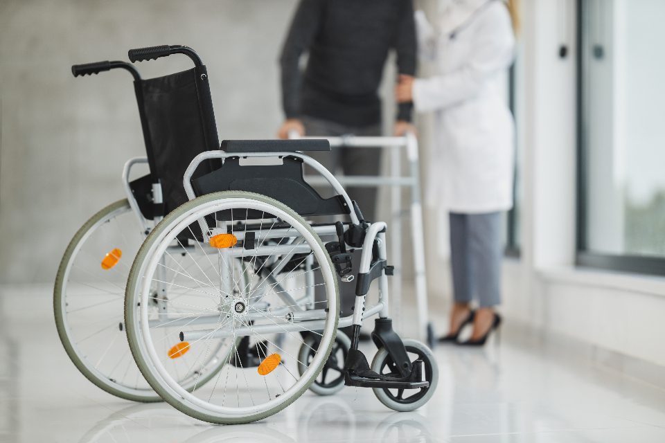 An empty wheelchair and a walking frame in a bright corridor, with two people out of frame