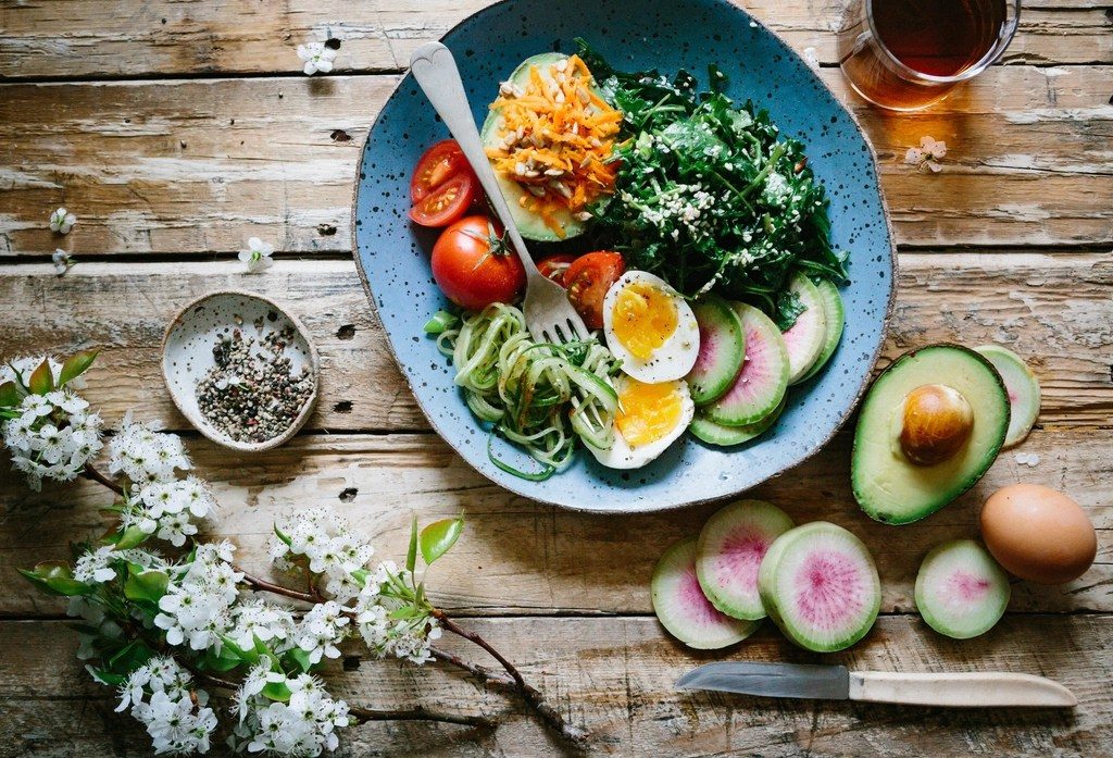 A blue bowl of vegetables, avocado and a poached egg on a wooden table