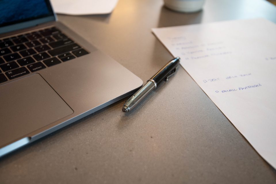 A laptop on a desk beside a pen and a sheet of handwritten notes