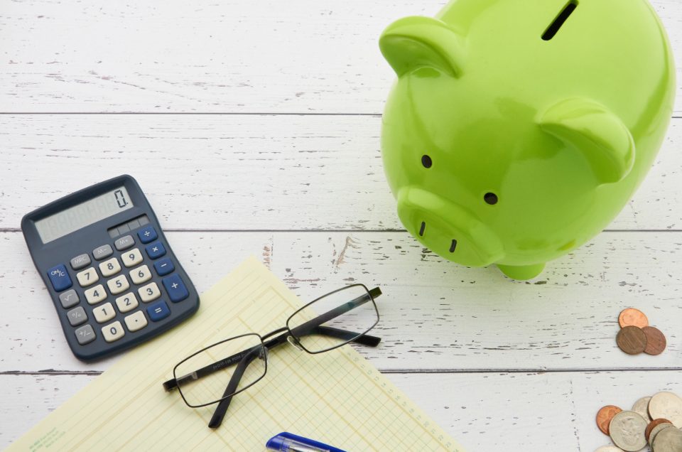 A green piggy bank beside a calculator, reading glasses and a few coins