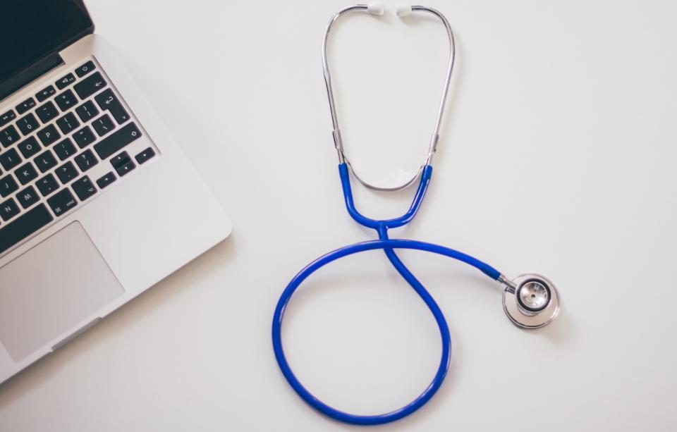 A blue stethoscope looped on a desk beside a laptop