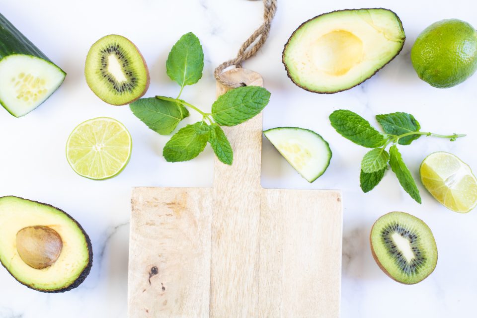 Halved avocados, kiwi fruit, limes, cucumber and mint around a wooden board